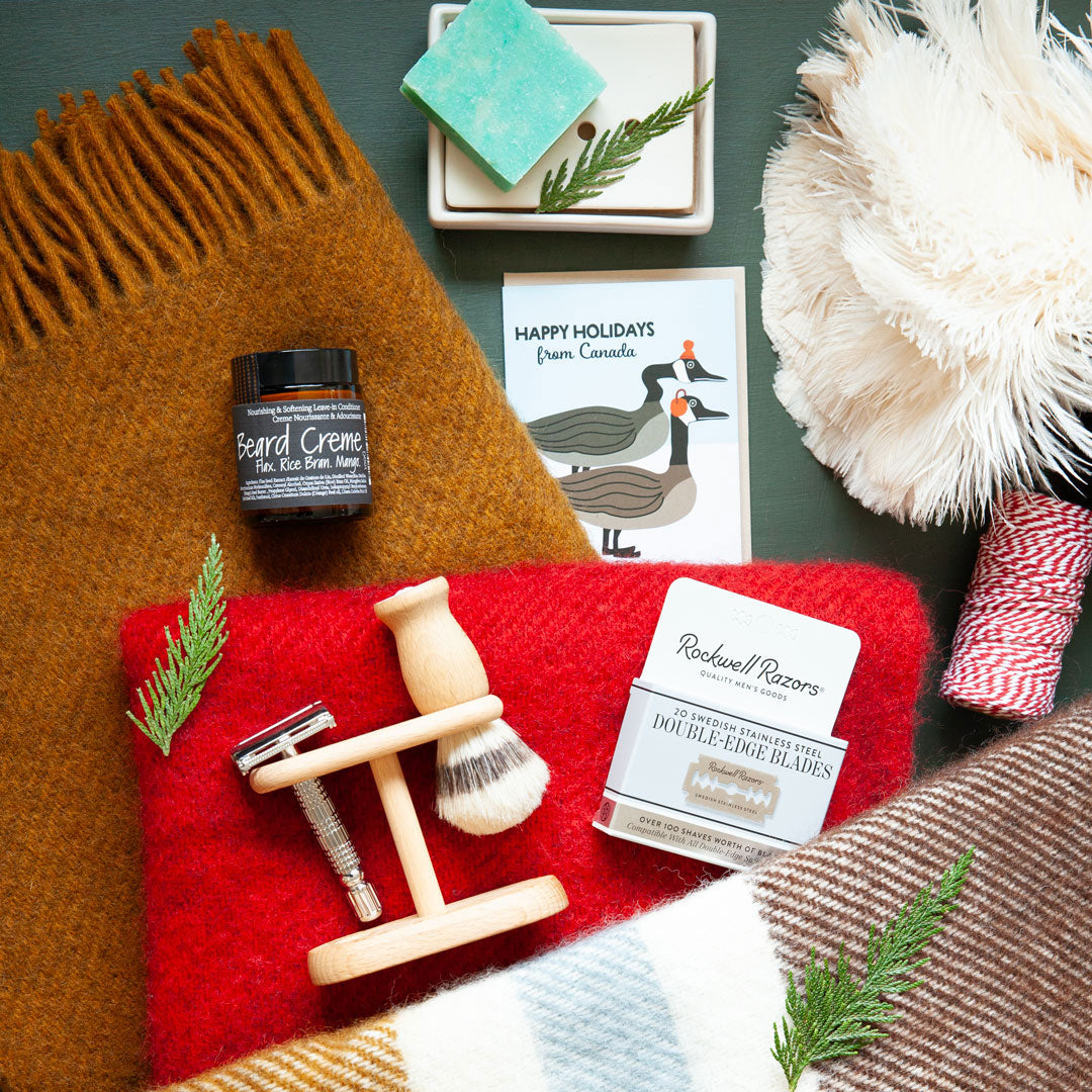 Grooming products including a jar of beard cream, shaving brush, and double-edge razor blades on a red blanket with festive decor.